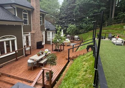 Wooden backyard deck with outdoor furniture, string lights, and a green lawn bordered by trees.