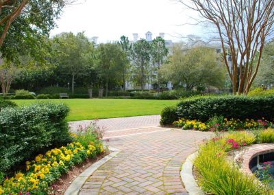 A garden with flower beds, brick paths, trees, and a grassy lawn with a building in the background.