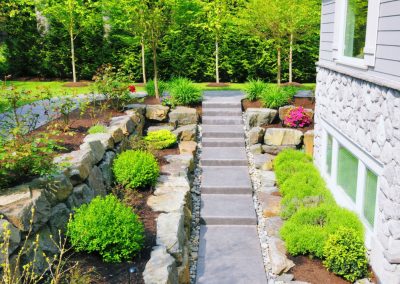 Stone steps bordered by shrubs and rocks lead to a modern house with a garden and lush green trees.