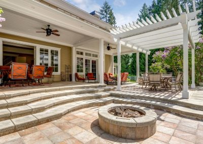 Sunny patio with steps, outdoor dining sets, a pergola, and a built-in fire pit surrounded by greenery.