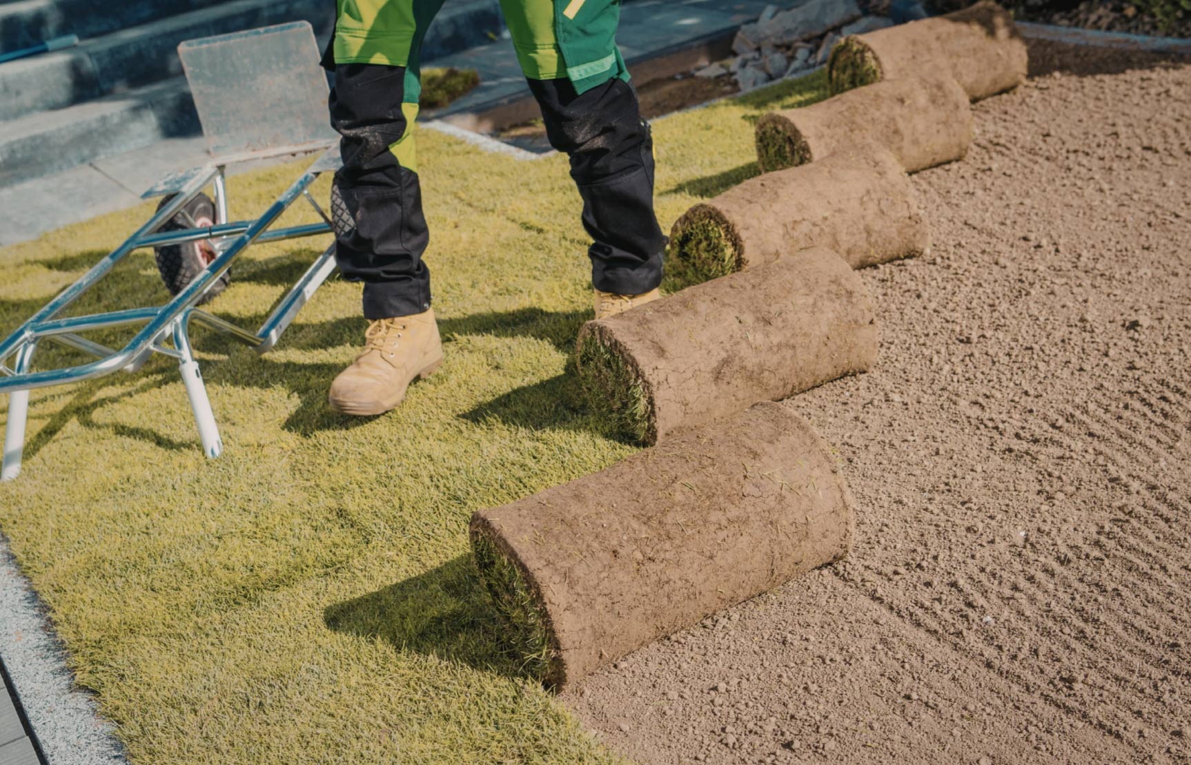 sod-installation-img Person laying rolls of sod on a yard with a wheelbarrow nearby and prepared soil visible.