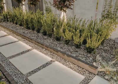 Stepping stones and gravel path beside neatly trimmed shrubs and a concrete wall in a landscaped garden.